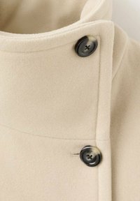 Kremowy płaszcz wykonany z miękkiej tkaniny, z wysokim kołnierzem i dwoma czarnymi guzikami z przodu, o gładkiej fakturze i prostych liniach.