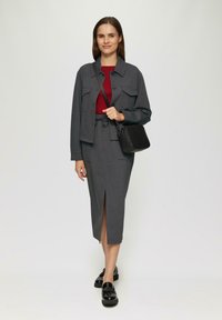 Grijze cropped jas met twee voorzakken, gecombineerd met een bijpassende grijze midi-rok met een split aan de voorkant, en zwarte schoenen, terwijl ze een kleine zwarte tas vasthoudt.