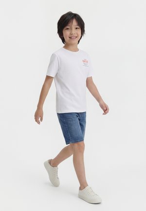 Garçon marchant, portant un t-shirt blanc à manches courtes avec logo, un short en jean et des baskets blanches, sur un fond blanc uni.
