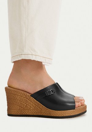 Piede con sandalo zeppa in pelle nera aperto sulle dita, suola marrone texturizzata e pantaloni bianchi corti.