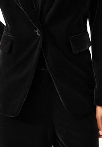 Czarny aksamitny żakiet o dopasowanym kroju, z klapami w kształcie notcha, dwoma przednimi kieszeniami i subtelnym blaskiem, zestawiony z pasującymi spodniami z aksamitu.