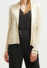 Ljusbeige kofta med öppen framdesign, långa ärmar och ett mjukt, något texturerat tyg, tillsammans med en svart outfit.