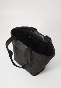 Czarna, pleciona torba na ramię z szerokimi uchwytami, skórzanym wykończeniem, otwartym wnętrzem i zamykaną na zamek kieszonką, stojąca na białej powierzchni.