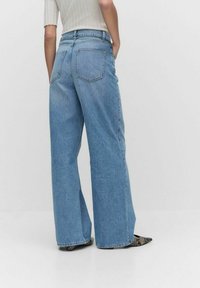 Vaaleansiniset leveälahkeiset farkut korkealla vyötäröllä, joissa on kaksi takataskua ja näkyvää ompelua, yhdistettyinä mustiin teksturoituihin kenkiin.