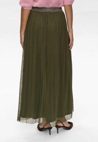 Olijfgroene plissé maxi-rok met een glanzende tailleband, doorschijnende stof en enkellange zoom. Draagt met bruin met hakken.