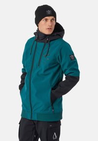 Giacca teal con accenti neri, tessuto resistente, cappuccio regolabile, cerniera frontale e dettagli logo sul petto e sul manica; abbinata a un berretto nero.