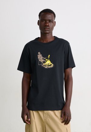 Camiseta negra de manga corta de algodón, con un estampado gráfico de un esqueleto junto a una fogata en tonos amarillos y grises.