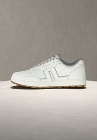 Λευκό sneaker χαμηλής κοπής με διάτρητη μύτη, γκρι επένδυση, κλείσιμο με κορδόνια και τακούνι από καφέ καουτσούκ πάνω σε απλή φωτεινή επιφάνεια.