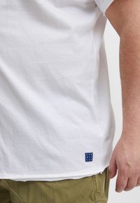 Mand iført hvid t-shirt med blåt firkantet logo og olivengrønne bukser, arm hvilende ned langs siden mod en ensfarvet baggrund.