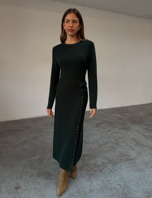 Vestido de malha verde escuro com mangas compridas, apresentando botões laterais e detalhes de destaque. Usado com botas bege de bico fino.