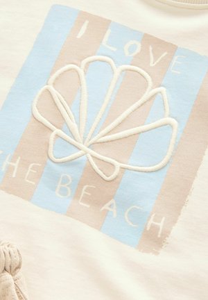 Tissu blanc avec un motif rayé bleu clair et beige, orné d'un coquillage brodé et du texte "J'AIME LA PLAGE" en blanc.