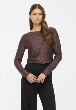 Un mannequin se tient devant un fond blanc uni, vêtu d’un haut à manches longues à motif floral couleur prune et d’un pantalon noir.
