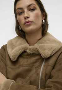 Giacca in finta suede marrone con colletto beige morbido, chiusura con zip e dettagli di cucitura visibili. Il tessuto appare morbido e testurizzato.
