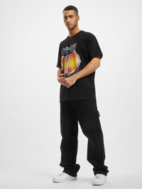 Svart grafisk t-skjorte med et fargerikt design på fronten, kombinert med løse svarte bukser og hvite sneakers. Enkle teksturer, casual passform.