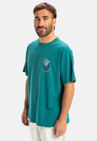 Camiseta de algodón en color verde azulado, con mangas cortas, escote redondeado y un diseño gráfico que presenta olas y un círculo en el pecho.