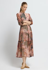 Dlouhá, patrová šaty z průhledné látky s paisley a geometrickými vzory v růžové, hnědé a černé barvě. Má vysoký krk a tříčtvrteční rukávy.