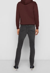Homme portant un jean slim gris foncé, des baskets noires et blanches, et un sweat à capuche bordeaux, debout de dos devant un fond uni.