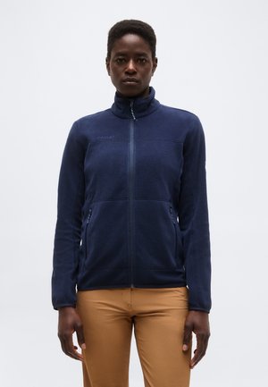 Γυναίκα που φοράει ζακέτα φλις σε σκούρο μπλε χρώμα με φερμουάρ και ανοιχτό καφέ παντελόνι, στέκεται μπροστά σε απλό ανοιχτό γκρι φόντο.