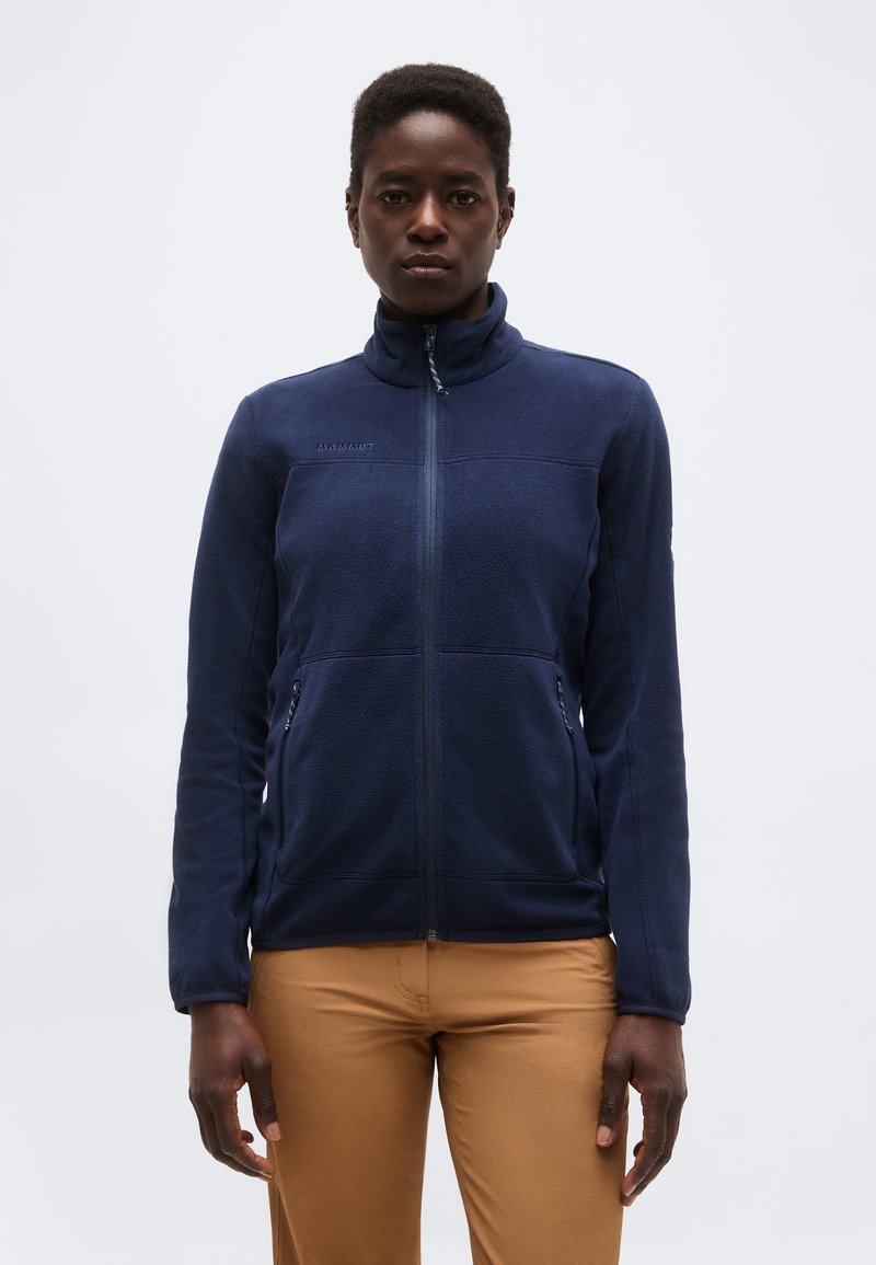 Nő sötétkék cipzáras polárdzsekiben és világosbarna nadrágban, egyszínű világosszürke háttér előtt állva.