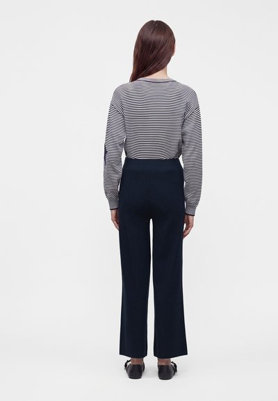 Personne debout de dos, portant une chemise à manches longues rayée noir et blanc, un pantalon large sombre et des chaussures noires.