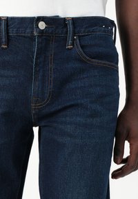 Jeans in denim blu scuro con cuciture arancioni a contrasto, cinque tasche, chiusura con bottone metallico e vestibilità dritta.