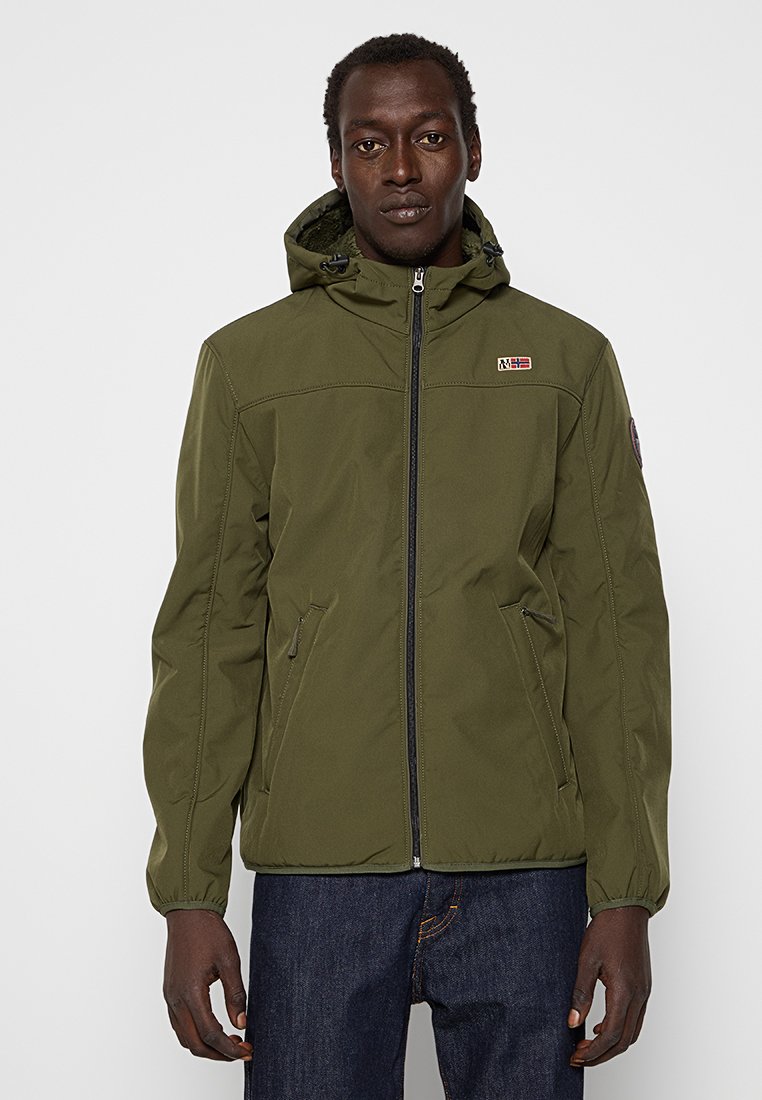 Hombre vestido con una chaqueta con capucha de color verde oliva y cremallera, con dos bolsillos laterales con cremallera, y jeans de color azul oscuro, frente a un fondo liso.