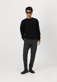Svart strikkegenser med frynsete kant, grå smale bukser og stilige svarte ankelstøvler. Modell med mørke solbriller. Enkel, moderne design.