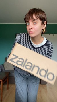 Kartono dėžė su ryškiais pilkais žodžiais "zalando". Pirmame plane matyti asmuo, vilkintis dryžuotą tamsiai mėlyną ir baltą marškinių, pilką kelnių.