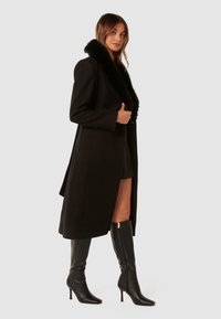 Abrigo de lana negro con cuello de piel sintética, con un corte largo y cierre de un solo botón, combinado con botas de cuero negras hasta la rodilla.