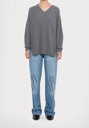 Personne portant un pull gris côtelé col en V et un jean bleu clair avec des logos AX bleu foncé sur l'avant des cuisses et des chaussures noires pointues.
