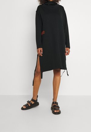 Schwarzes, langärmeliges Kleid mit hohem Kragen, seitlichen Schlitzen und einem Akzent in Form roten Textes auf der einen Seite. Hergestellt aus einem weichen Stoff mit lockerer Passform.