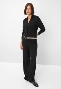Zwarte cardigan met een V-hals, knoopsluiting en contrasterende crème strepen. Gecombineerd met wijde zwarte broek en zwarte schoenen.