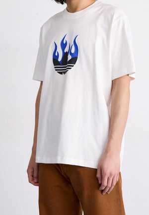 Person trägt ein weißes T-Shirt mit einem blau-schwarzen Flammenlogo auf der Brust und braune Hose vor neutralem Hintergrund.