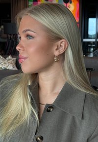 Guldörhängen med en vågig design, bärs av en person med långt blont hår, olivgrön jacka och ett fint kedjearmband.