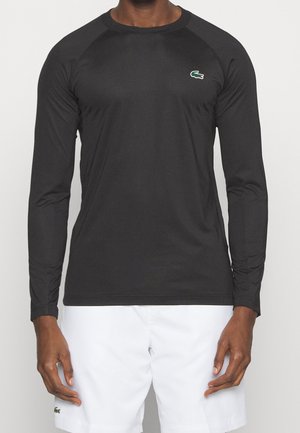 Mann trägt schwarzes langärmeliges Sportshirt mit grünem Krokodil-Logo und weißen Shorts mit passendem Logo, steht vor neutralem Hintergrund.