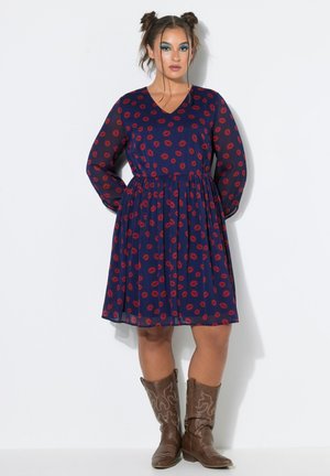 Frau steht mit den Händen hinter dem Rücken, trägt ein dunkelblaues Kleid mit rotem Lippen-Print und braune Cowboy-Stiefel vor weißem Hintergrund.
