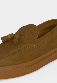 Mocassin en daim marron avec des glands sur le dessus et une semelle en caoutchouc épaisse assortie sur un fond clair uni.