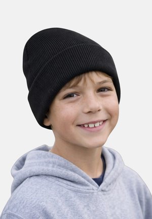 Lächelnder Junge mit schwarzer Strickmütze und hellgrauem Hoodie, der leicht zur Seite schaut vor einem einfarbigen Hintergrund.