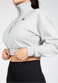 Lichtgrijze cropped sweatshirt met een hoge kraag, rits en zwart logo. Bevat ribgebreide manchetten en een gladde textuur. Gecombineerd met zwarte leggings.