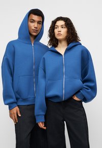 Μπλε φούτερ με φερμουάρ, ευρύχωρες τσέπες, φαρδιά μανίκια και άνετη εφαρμογή. Κατασκευασμένα από μαλακό υλικό, παρουσιάζονται σε δύο μοντέλα μπροστά σε ουδέτερο φόντο.