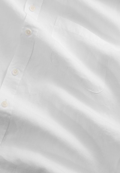 Gros plan sur l'avant d'une chemise blanche en lin montrant les boutons et la poche poitrine avec une texture de tissu visible et des ombres douces.