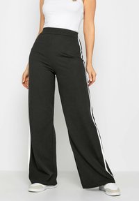 Pantalons larges noirs avec des rayures blanches sur les côtés, fabriqués en tissu doux. La taille haute crée une silhouette décontractée. Associés à des baskets blanches.