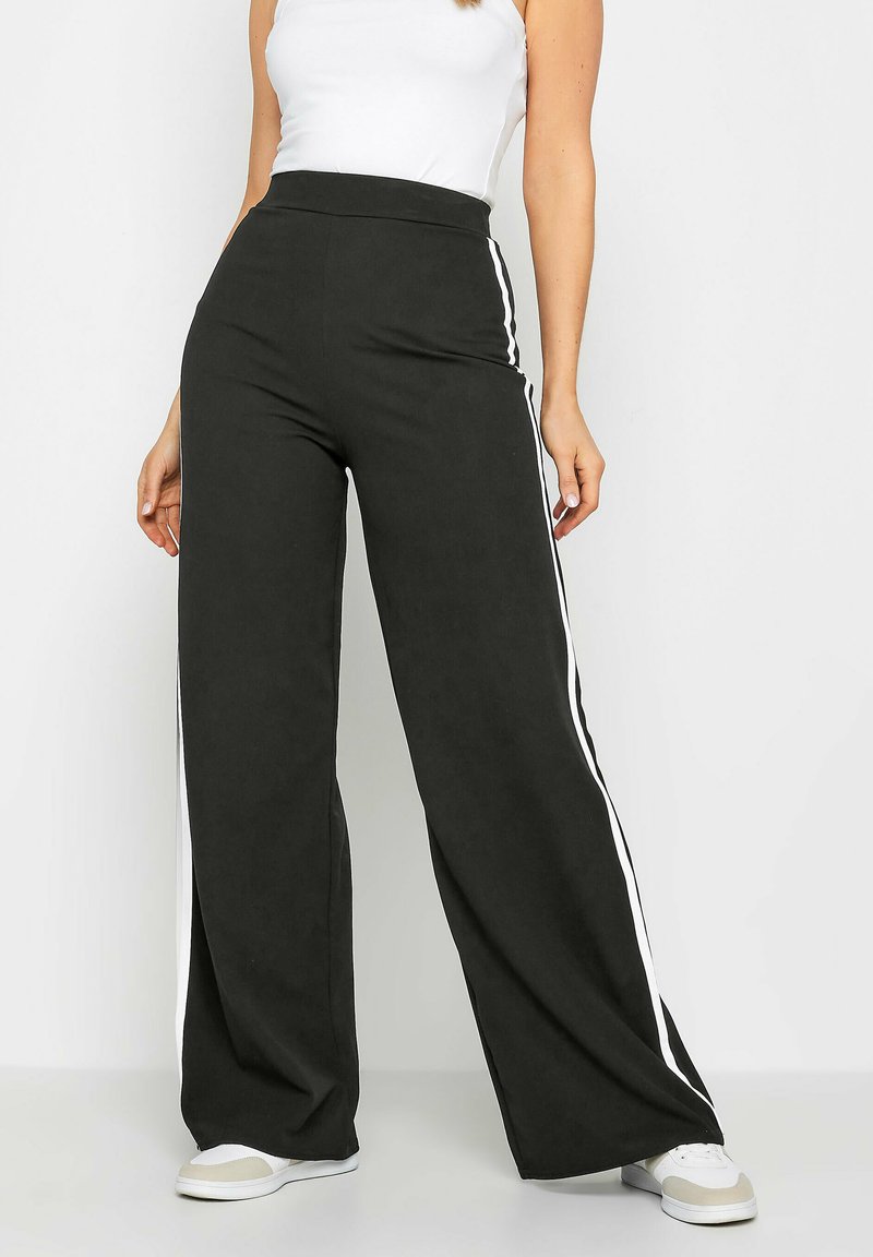 Pantalons larges noirs avec des rayures blanches sur les côtés, fabriqués en tissu doux. La taille haute crée une silhouette décontractée. Associés à des baskets blanches.