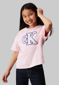T-shirt in cotone rosa con grande ricamo "cK" in blu, maniche corte, vestibilità rilassata, abbinato a jeans in denim scuro.