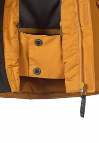 Isolerad jackas insida i klart orange, med en zip-ficka och knappstängning, samt en svart tygutsida och gula detaljer.