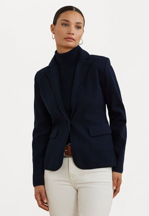 Marinblå ullblazer med skräddarsydd passform, bröstfickor med utskärning, två framfickor och knapp i mitten. Modellen har den över en polotröja.