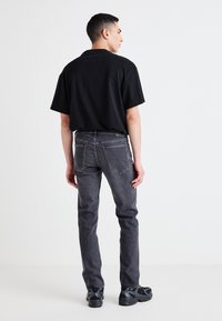 T-shirt en coton noir associé à un jean gris foncé. Le jean présente des poches arrière et un discrète étiquette de logo. La chaussure est des sneakers noires.