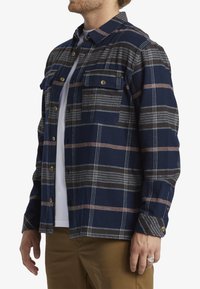 Camisa de franela a cuadros azul marino con rayas blancas y naranja quemado, que presenta dos bolsillos frontales y acentos metálicos en los botones.