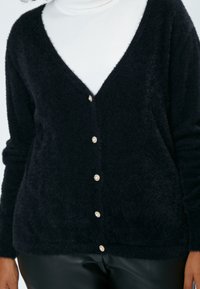 Schwarze, flauschige Strickjacke mit tiefem V-Ausschnitt und perlenartigen Knöpfen auf der Vorderseite, getragen über einem weißen Rollkragenpullover. Strukturiertes Material.
