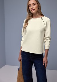 Femme portant un haut texturé crème à manches longues avec un détail zippé et un jean bleu foncé, tenant un sac en daim marron avec une doublure à motifs.